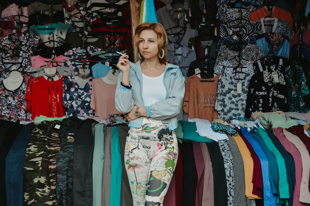 Thoughtful female in casual clothes leaning on shelf with garments while working in market and smoking cigarette